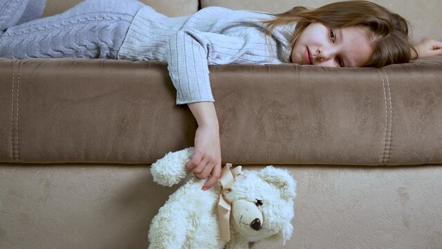 Sad girl hugging teddy bear lying on sofa