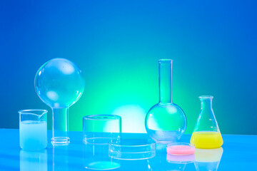 Front view of boiling flasks, erlenmeyer flask, beaker and petri dish containing color liquid decorated on blue gradient background. Space on glass podium for product presentation