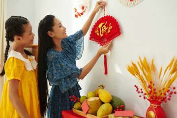 Cheerful mother and teenage daughter hanging traditional spring festival decorations on apartment...