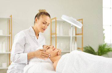 Fototapeta premium Professional female beautician or cosmetologist examining skin condition to a young pretty girl client with special lamp in beauty spa salon. Facial massage, skin care and treatment concept.