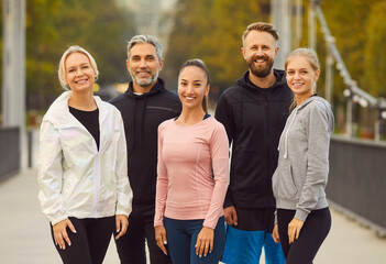 Diverse friends enjoy active outdoor fitness training workouts. Group portrait happy young and mature multiethnic men and women in sports clothes standing on city bridge, looking at camera and smiling