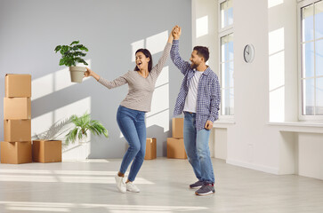 Happy young couple celebrating moving day with cardboard boxes in background smiling and dancing with plant in hands in new appartment. Family having fun. Relocating, real estate, mortgage concept.