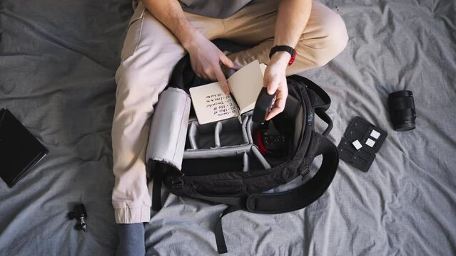 A professional photographer collects equipment in a special backpack, noting the items in a list in a notebook. Flat layout top view.