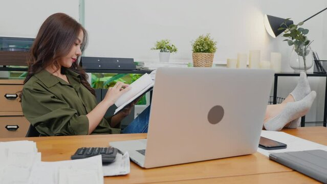 Asian young businesswoman sitting relaxing reclining with feet up on computer desk reading report document in paperwork on clipboard, smile woman holding document on hand reading letter at home office