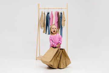 Obraz premium A child, a little girl, stands near the closet, chooses clothes against a light background. Dressing room with clothes on hangers. Wardrobe of children's and stylish clothes. Montessori wardrobe.