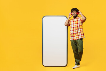 Full body young happy Indian man he wears shirt casual clothes big huge blank screen mobile cell phone smartphone with mockup area listen music in headphones isolated on plain yellow color background.