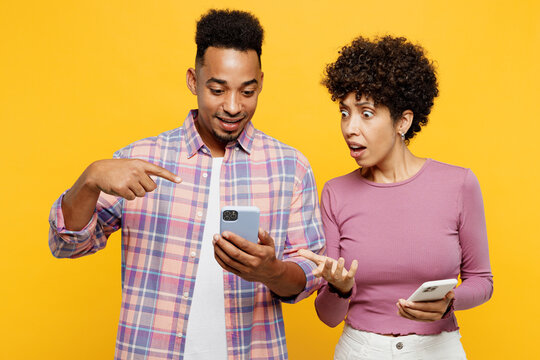 Young Shocked Sad Couple Two Friends Family Man Woman Of African American Ethnicity Wearing Purple Casual Clothes Together Hold Use Show Mobile Cell Phone Isolated On Plain Yellow Orange Background.