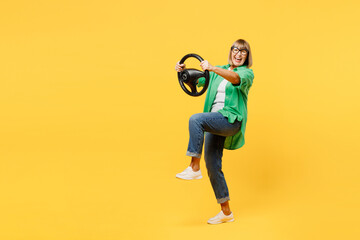 Full body side view elderly blonde woman 50s years old wear green shirt glasses casual clothes hold steering wheel driving car isolated on plain yellow background studio portrait. Lifestyle concept.