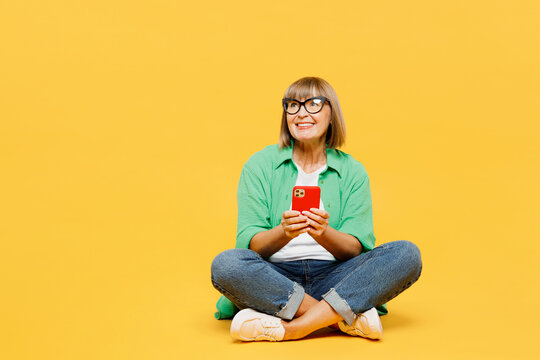 Full Body Elderly Blonde Woman 50s Year Old Wear Green Shirt Glasses Casual Clothes Sitting Hold In Hand Use Mobile Cell Phone Look Aside On Area Isolated On Plain Yellow Background Lifestyle Concept