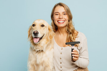 Young fun owner woman wear casual clothes hug cuddle embrace best friend retriever dog hold grooming brush isolated on plain pastel light blue background studio portrait. Take care about pet concept.