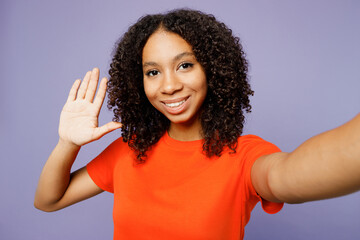 Close up little kid teen girl of African American ethnicity wear orange t-shirt do selfie shot mobile cell phone wave hand greet isolated on plain pastel purple background Childhood lifestyle concept