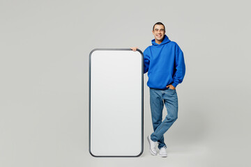 Full body happy young middle eastern man he wear blue hoody casual clothes big huge blank screen mobile cell phone with workspace copy space area look camera isolated on plain solid white background.