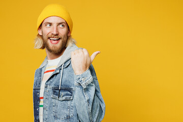 Young smiling happy fun blond man he wears denim shirt hoody beanie hat casual clothes point thumb finger aside on area mockup isolated on plain yellow background studio portrait. Lifestyle concept.