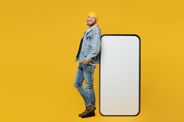 Full body side view young man wear denim shirt hoody beanie hat casual clothes stand near big huge blank screen area mobile cell phone smartphone isolated on plain yellow background Lifestyle concept - Powered by Adobe
