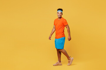 Full body side view happy young fitness trainer sporty man sportsman wear orange t-shirt walk go look camera spend time in home gym isolated on plain yellow background. Workout sport fit abs concept. - obrazy, fototapety, plakaty