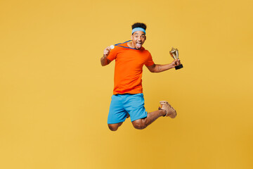 Fototapeta na wymiar Full body young fitness trainer instructor sporty man sportsman wear orange t-shirt jump high golden medal champion cup award in gym isolated on plain yellow background. Workout sport fit abs concept.