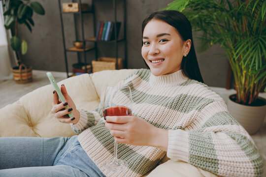 Young cheerful happy woman wear casual clothes drink red wine use mobile cell phone sits on armchair stay at home hotel flat rest relax spend free spare time in grey living room indoor Lounge concept