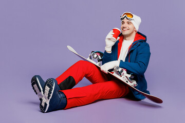 Full body minded smiling man wear blue windbreaker jacket ski goggles mask hat sitting hold snowboard drink coffee spend extreme weekend winter season in mountains isolated on plain purple background.