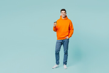 Full body young man he wears orange hoody casual clothes hold takeaway delivery craft paper brown cup coffee to go isolated on plain pastel light blue cyan color background studio. Lifestyle concept.