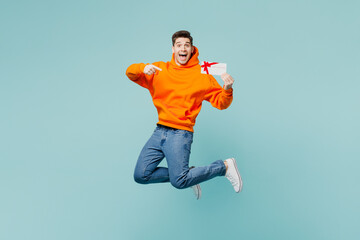 Full body young man he wears orange hoody casual clothes jump high hold point finger on gift certificate coupon voucher card for store isolated on plain blue cyan color background. Lifestyle concept. - obrazy, fototapety, plakaty