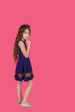 Full length of elegance cover girl in blue dress with index finger to lips at pink isolated, looking at camera. Emotional kid lady 9 year old, studio shot. Child emotion concept. Copy ad text space