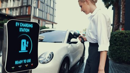 Businesswoman pull out EV charger from charging station to recharge her electric car at city residential building. Modern urban lifestyle and eco-friendly electric car utilization to reduce CO2.Peruse