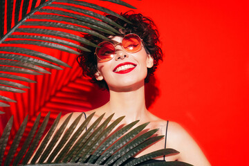 Fashion beauty portrait of young smiling brunette woman with evening stylish  makeup. Sexy model with curly short hair posing in studio. With red bright lips. Tropical palm leaf. In sunglasses