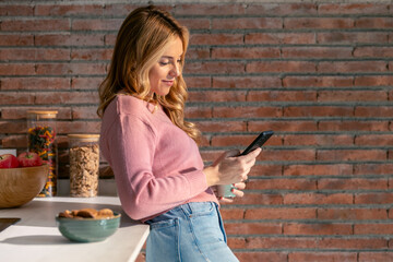 Smiling young woman using her mobile phone while drinking a cup of coffee in the kitchen at home.