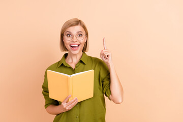 Portrait of genius overjoyed girl wear khaki shirt in eyewear hold book raising finger up have idea isolated on pastel color background