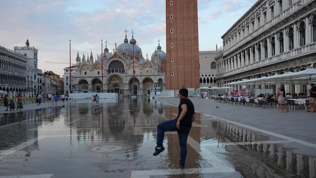 Young man splashing in water due to high tide in St. Mark's Square in front of Campanile and Basilica in Venice, Italy