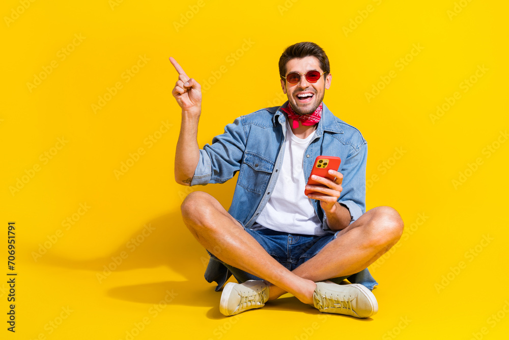 Poster full size photo of pleasant guy wear jeans jacket sit hold smartphone indicating at offer empty spac