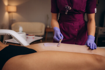A master applies pink depilatory wax to a young woman's leg for hair removal. Depilation with wax. Beauty concept. Place for text. Selective focus. - obrazy, fototapety, plakaty