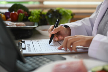 Side view of nutritionist writing diet plan and using laptop at desk with different healthy products
