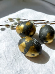 Dark blue and gold Easter eggs lie on a white light tablecloth under the rays of the sun, willow branches lie nearby. An Easter card. Vertical photo