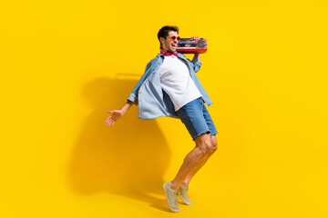Fototapeta na wymiar Full size photo of eccentric guy wear jeans jacket in sunglass boombox on shoulder look empty space isolated on yellow color background
