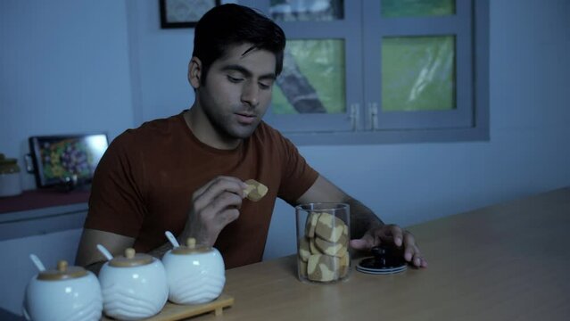 Indian man satisfying his sugar craving - eating cookies at night  midnight craving  late-night snacking  eating disorder. Attractive youth enjoying homemade biscuits - unhealthy food  diet fail  g...