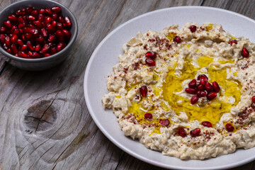 Moutabal - smoky aubergine with a touch of tahini and garlic on a plate topped with pomegranate arils, sumac, and olive oil. A bowl with pomegranate arils on the side.