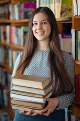 Portrait of a female student with a stack of books in library. Cheerful caucasian girl studying for college exams, prepares class assignment