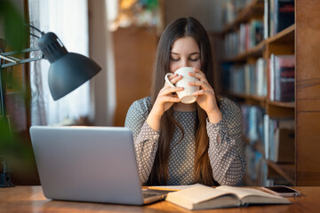Girl student having a break, drinking tea or coffee sitting at desk with books and laptop. Student...
