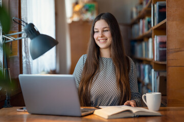 Fototapeta premium Smiling girl having online lesson via the Internet. Cheerful female student in university library, preparing assignment