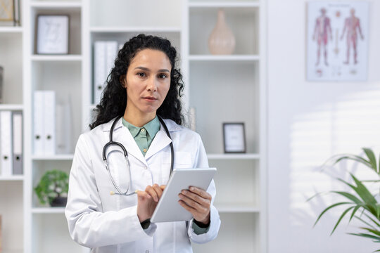 Close-up portrait of young female doctor, Latin American woman using tablet computer, looking serious pensive and focused at camera, standing inside office in medical office of clinic.