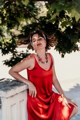 woman in a red silk dress and a bandage on her head smiles against the background of the leaves of a tree. She is leaning on the coop and looking into the camera. Vertical photo.