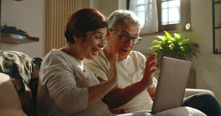 At home, a couple of grandparents make a video call to their relatives and friends using the smartphone . The elderly couple smiles, blows kisses and is happy to talk to them. - Powered by Adobe