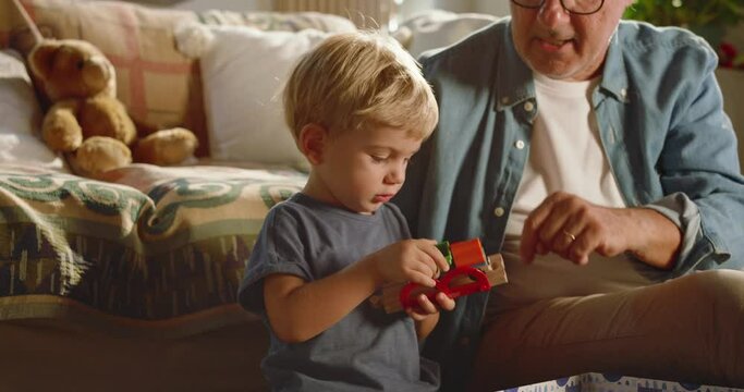 At home, at sunset, the grandfather and his beautiful grandson play with a wooden train and teaches him to build the track to play while they have fun
