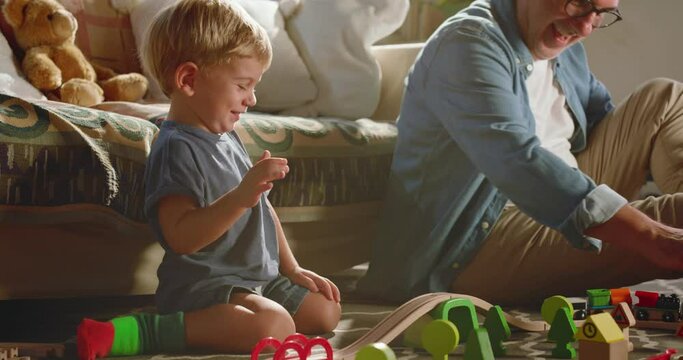 At home, at sunset, the grandfather and his beautiful grandson play with a wooden train and teaches him to build the track to play while they have fun