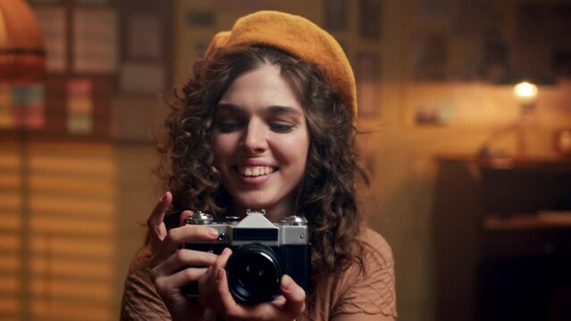 Pretty young woman in yellow beret smiles and takes photos with vintage camera indoors. girl in retro clothes takes photographs in old office or studio. Portrait of attractive photographer.