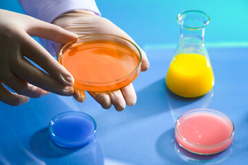 Hand model wearing medical gloves with a glass petri dish of orange chemical liquid. Liquid in...