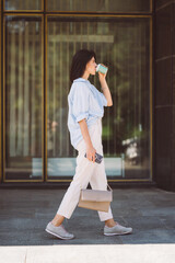 Full body shot of a confident young woman walking outdoors and drinking coffee.