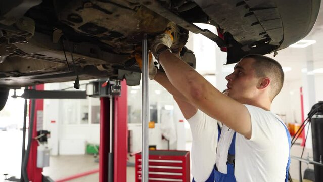 Auto mechanics in uniform install gearbox, repair car clutch in modern workshop. Professionals work on vehicle transmission maintenance with tools, demonstrate mechanical skills under raised auto.