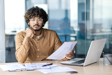 Portrait of serious concentrated businessman inside office, man looking at camera holding papers financial reports, accountant financier with laptop on paper work.